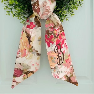 May include: A floral patterned scarf with a brown monogram letter "B" is displayed against a green wreath and a light blue background. The scarf features a watercolor-style design with pink, white, and brown hues.