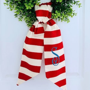 May include: Red and white striped fabric bow with a blue embroidered letter "S" on the right side. The bow is tied around a green wreath.
