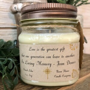 May include: A glass jar candle with a brown lid and a kraft paper label. The label has a gold star design and the text "Love is the greatest gift that one generation can leave to another. In Loving Memory - Jean Demos". The candle is labeled "French Lilac" and "8 oz. Soy". The label also includes the text "Risen Flame Candle Company".