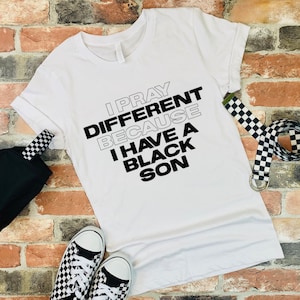 May include: White t-shirt with the text "I PRAY DIFFERENT BECAUSE I HAVE A BLACK SON" in black and gray. The shirt is paired with black and white checkered sneakers, a matching belt, and a black bag. The items are arranged on a brick wall.