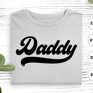 K&ouml;nnte beinhalten: Ein wei&szlig;es T-Shirt mit dem Wort "Daddy" in einer schwarzen Retro-Schrift.