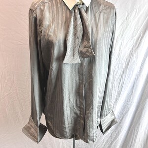 May include: A gray, long-sleeved blouse with a vertical striped pattern. The blouse features a white collar and a matching gray tie. The cuffs are buttoned. The blouse is displayed on a mannequin.