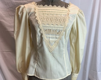 May include: A cream-colored blouse with long sleeves and a lace overlay on the front and cuffs. The lace is a delicate white and has a floral pattern.