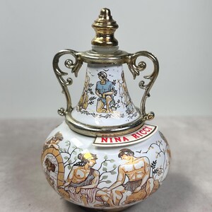May include: A white ceramic perfume bottle with a rounded base and a tapered neck featuring gold-tone metal handles and a matching stopper. The surface is decorated with colorful illustrations of figures in classical attire and the text NINA RICCI.