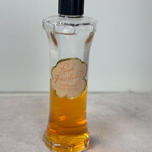 May include: A vintage cologne bottle with a black cap and a clear glass body. The bottle contains a golden-colored liquid and features a label that reads "Pink Frosting Cologne". The bottle has a unique, hourglass-like shape.