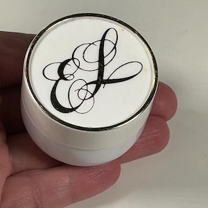 May include: A small, white container with a silver rim. The lid features a white background with the black cursive letters "E" and "L" intertwined. The container is held in a hand.