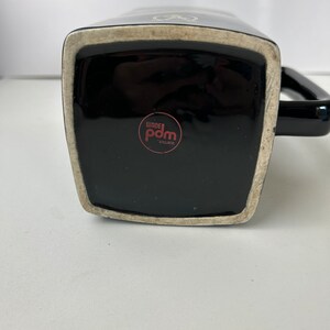 K&ouml;nnte beinhalten: Schwarze Keramiktasse mit wei&szlig;em Design und schwarzem Henkel. Der Boden der Tasse hat einen roten Kreis mit dem Text "Wade Pdm England".