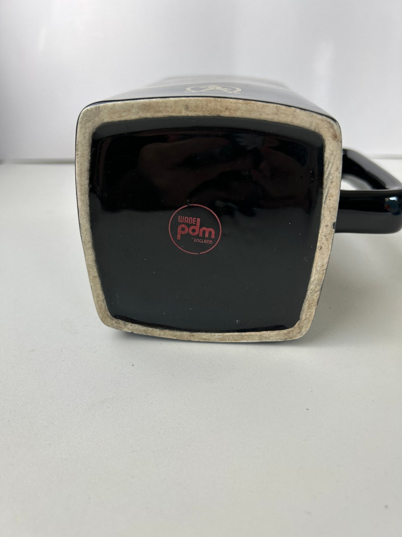 K&ouml;nnte beinhalten: Schwarze Keramiktasse mit wei&szlig;em Design und schwarzem Henkel. Der Boden der Tasse hat einen roten Kreis mit dem Text "Wade Pdm England".