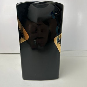 K&ouml;nnte beinhalten: Schwarze Keramikvase mit goldenen geometrischen Mustern. Die Vase hat eine rechteckige Form mit leicht abgerundeter Oberseite.