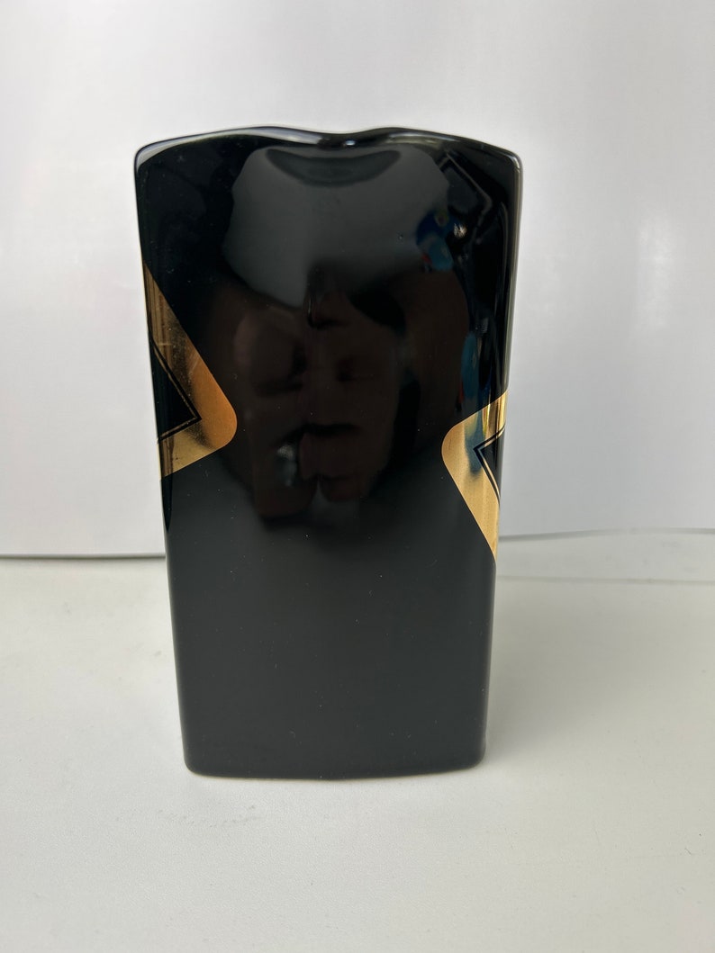 K&ouml;nnte beinhalten: Schwarze Keramikvase mit goldenen geometrischen Mustern. Die Vase hat eine rechteckige Form mit leicht abgerundeter Oberseite.