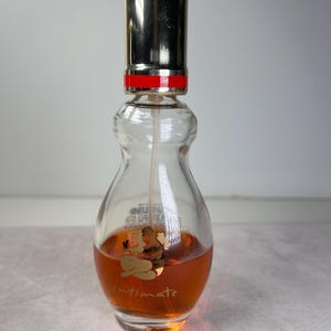May include: A clear glass perfume bottle with a silver cap and a red band. The bottle contains a light brown liquid and features a gold label with the word "Intimate" and a graphic. The bottle has a unique, curvy shape.