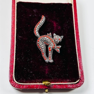 May include: A silver and red cat brooch in a red velvet box. The cat is made of silver and has red enamel accents. The brooch is in a red velvet box with a gold clasp.