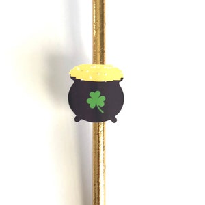May include: A gold paper straw with a St. Patrick's Day themed decoration. The decoration is a black pot of gold with a green shamrock and a yellow lid. The straw is a festive accessory.