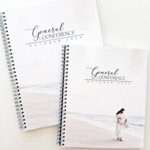 May include: Two spiral-bound notebooks with white covers featuring a beach scene and the text General Conference October 2026. The smaller notebook displays a figure in white robes carrying a child on a sandy shore by the ocean.