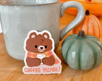 Puede incluir: Un oso marrón con una bufanda roja y naranja sostiene una taza de café. La pegatina dice "Coffee Please!" y está sobre una superficie de madera con una taza gris y calabazas verdes y naranjas.