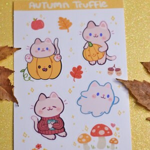 May include: A sheet of stickers featuring cute cartoon cats in various autumnal scenes. The stickers include cats wearing pumpkins, holding pumpkins, wearing sweaters, and flying as a ghost. The sheet also includes mushrooms, acorns, and leaves. The text "Autumn Truffle" is at the top of the sheet and "@PlanetBunnie" is at the bottom.