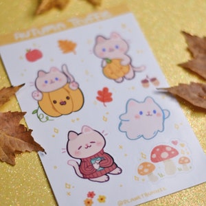 May include: A sheet of stickers featuring cute cartoon cats in various autumnal scenes. The stickers include a cat holding a pumpkin, a cat wearing a sweater, a cat with a mushroom, and a cat with a leaf. The stickers are on a white background with a light yellow border. The text "Autumn Treats" is at the top of the sheet.