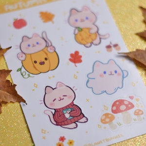 May include: A sheet of stickers featuring cute cartoon cats in various autumnal scenes. The stickers include cats holding pumpkins, wearing sweaters, and floating in clouds. There are also mushrooms and leaves. The sheet is white with a light pink background and the text "Autumn Treats" at the top.