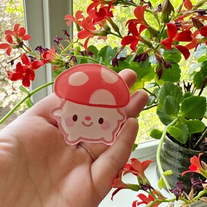 May include: A pink and red mushroom-shaped sticker with a cute cartoon face. The sticker is held in a hand against a backdrop of green foliage and red flowers.