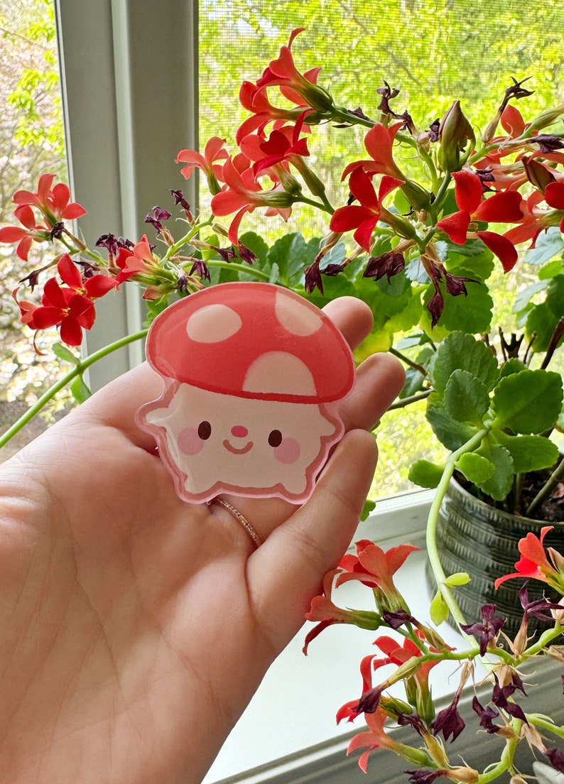 May include: A pink and red mushroom-shaped sticker with a cute cartoon face. The sticker is held in a hand against a backdrop of green foliage and red flowers.