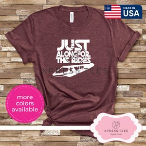 May include: Maroon t-shirt with the text "JUST ALONG FOR THE RIDES" in white, along with a white graphic of a ride vehicle. The shirt also has a "MADE IN USA" tag and a pink circle that says "more colors available".
