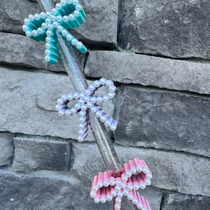 May include: Three decorative drink stirrers with bow accents. The bows are in shades of teal, lavender, and pink, each adorned with white pearl-like beads. The stirrers are clear with glitter accents, set against a gray stone background.