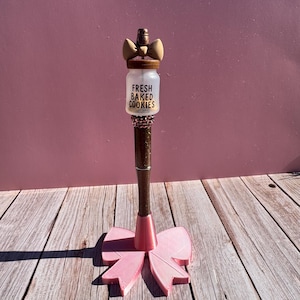 May include: A decorative pen with a pink bow-shaped base and a brown shaft. The pen features a small jar with the words "FRESH BAKED COOKIES" and a bow on top. The pen is set against a pink background and wooden surface.