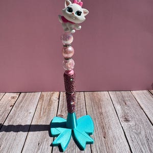 May include: A decorative pen featuring a white cat figurine with a pink bow and collar. The pen has a pink glitter barrel, pink and clear beads, and a turquoise bow-shaped base. The pen is approximately 7 inches tall.