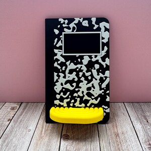 May include: A rectangular black and white composition notebook with a speckled pattern and a blank white-bordered label rectangle at the top. A bright yellow, semi-circular, ribbed shelf is attached to the lower front of the notebook cover.