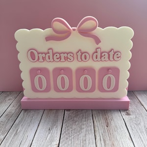 May include: A pink and white decorative sign with the text "Orders to date" and the number "0000". The sign has a scalloped edge and a bow at the top. The sign is on a pink base.