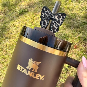 Puede incluir: Un vaso Stanley marrón con borde dorado y el logotipo de Stanley. Un lazo con estampado de leopardo en blanco y negro está sujeto a la pajita. El vaso lo sostiene una persona con esmalte de uñas rosa.