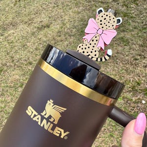 Könnte beinhalten: Braune Stanley-Reisebecher mit goldenen Akzenten und einem schwarzen Deckel. Eine dekorative Leopardfigur mit rosa Schleife sitzt oben. Der Becher wird von einer Person mit rosa Nagellack gehalten.