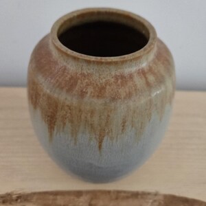 Pode incluir: Um pequeno vaso redondo de cerâmica com um esmalte azul claro e detalhes marrons. O vaso tem uma abertura larga e um fundo arredondado.