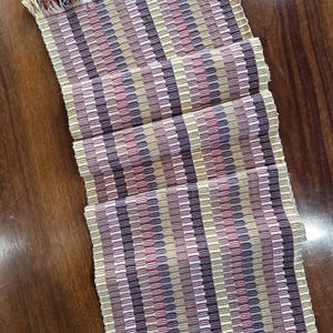 Puede incluir: Un camino de mesa rectangular tejido con un patrón de rayas en tonos beige, marrón, rojo y amarillo. El camino de mesa tiene bordes con flecos y se muestra sobre una superficie de madera.