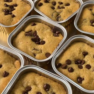 Może przedstawiać: Zbliżenie na kilka kwadratowych ciasteczek z kawałkami czekolady w indywidualnych pojemnikach z folii. Ciasteczka są złocistobrązowe z rozrzuconymi kawałkami czekolady. Pojemniki są srebrne i blisko siebie.