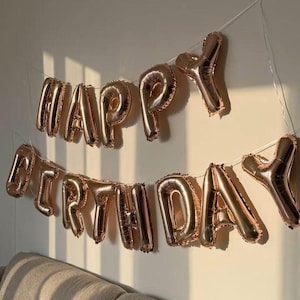 May include: A set of rose gold foil balloons spelling out the words HAPPY BIRTHDAY, arranged in two rows on a string. The metallic balloons have a reflective surface and are suspended against a plain wall, catching the natural light.