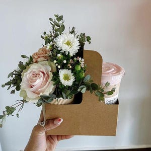 Pode incluir: Um suporte de cartão castanho contém um buquê de flores e uma bebida. O buquê inclui uma rosa rosa claro, crisântemos brancos e vegetação. A bebida está em um copo de plástico transparente com tampa rosa.