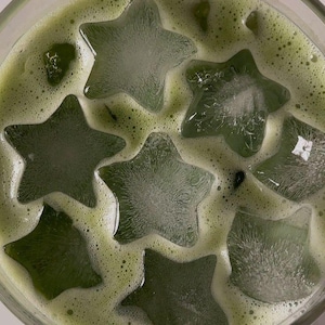 May include: A refreshing green beverage with star-shaped ice cubes. The drink has a frothy texture and a light green color, with the ice cubes adding a unique visual element. The drink is served in a clear glass.