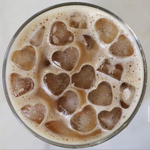 Könnte beinhalten: Ein erfrischender Eiskaffee in einem klaren Glas, gefüllt mit herzförmigen Eiswürfeln. Der Kaffee hat eine hellbraune Farbe, und das Getränk ist mit einer schaumigen, cremigen Schicht bedeckt. Das Glas steht vor einem weißen Hintergrund.