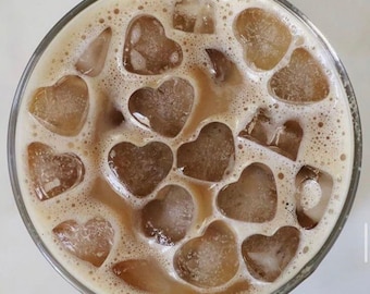 May include: A refreshing iced coffee drink in a clear glass, filled with heart-shaped ice cubes. The coffee is a light brown color, and the drink is topped with a foamy, creamy layer. The glass is set against a white background.