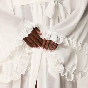 May include: A white robe with ruffled sleeves and a matching tie belt. The robe is made of a lightweight, flowing fabric. The sleeves and edges of the robe are trimmed with delicate ruffles. The robe is tied at the waist with a matching belt.
