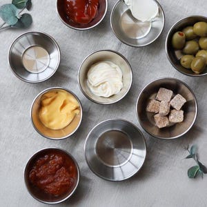 Puede incluir: Una colección de pequeños cuencos de acero inoxidable llenos de diversos condimentos e ingredientes, como ketchup, mayonesa, aceitunas y terrones de azúcar. Los cuencos están dispuestos sobre una superficie gris claro, para servir.