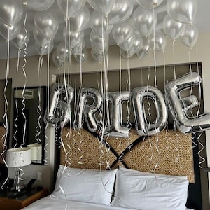 Puede incluir: Una habitación decorada para una celebración con globos plateados y una gran exhibición de globos con la palabra "BRIDE". Cintas y serpentinas blancas cuelgan del techo. La cama está hecha con sábanas y almohadas blancas, con un cabecero estampado.