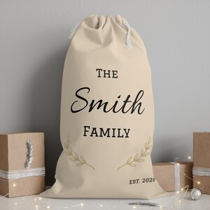 May include: A beige drawstring sack featuring the black printed text THE Smith FAMILY and EST. 2026. The design includes two gold-coloured leaf sprigs flanking the text. The bag is made of fabric and has a white cord closure at the top opening.