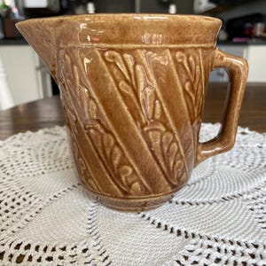 May include: A brown ceramic pitcher with a spout and handle. The pitcher has a textured, diagonal leaf pattern. It sits on a white, crocheted doily. The pitcher is likely vintage or antique.