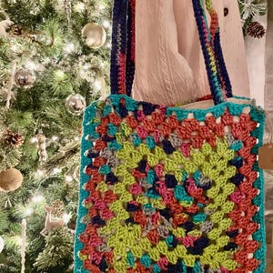 May include: A colorful, handmade crochet tote bag. The bag features a square granny square design in shades of teal, pink, green, and blue. The straps are a mix of rainbow colors. The bag is displayed in front of a Christmas tree.