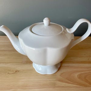 May include: A white ceramic teapot with a lid and handle. The teapot has a rounded body with a scalloped edge and sits on a small pedestal base. The spout and handle curve gracefully. The teapot is on a wooden surface.