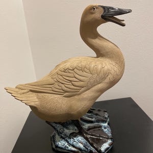 May include: A tan ceramic duck sculpture with a black beak and open mouth. The duck is perched on a dark, textured base with blue and white accents. The sculpture has detailed feather and wing patterns.