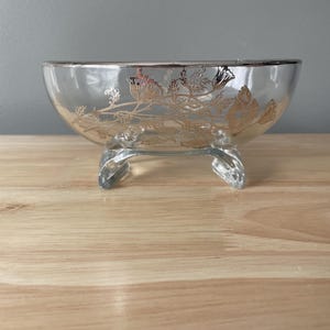 May include: A clear glass bowl with a silver floral design and three legs. The bowl is on a wooden surface.