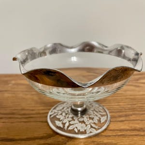 May include: A clear glass dessert dish with a silver-colored rim and base. The rim has a wavy design. The base features a floral pattern. The dish is elevated on a short stem.